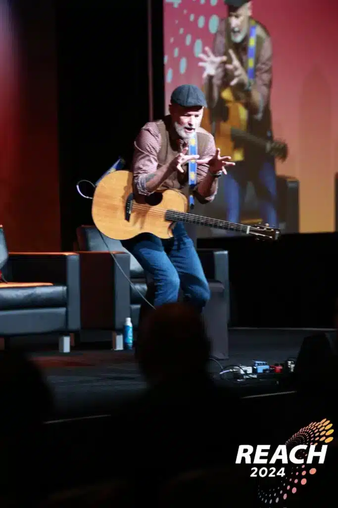 Mike Rayburn speaking at REACH 2024, engaging the audience with guitar-driven keynote full of insight, energy, and entertainment